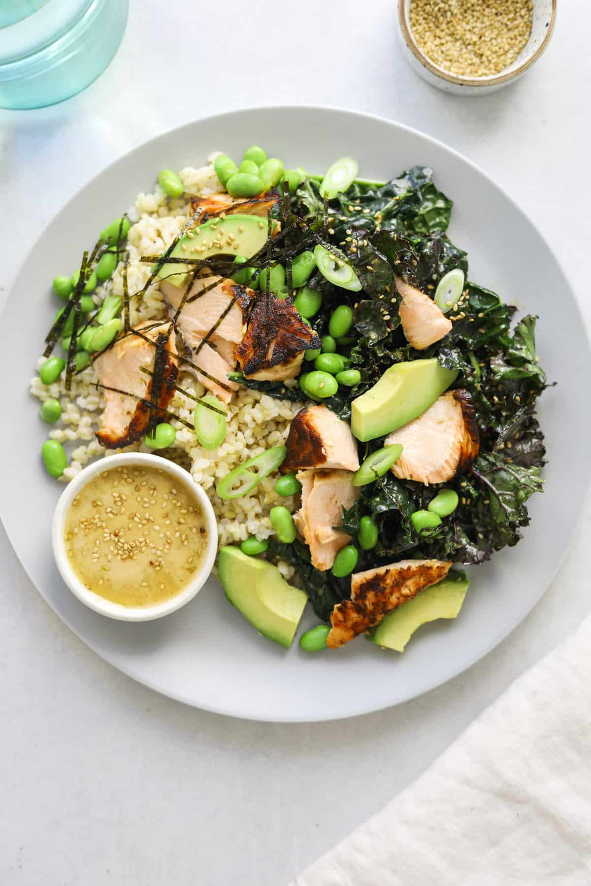 a close up of a plate of brown rice, salmon and kale salad with a small side bowl of miso dressing topped with toasted sesame seeds