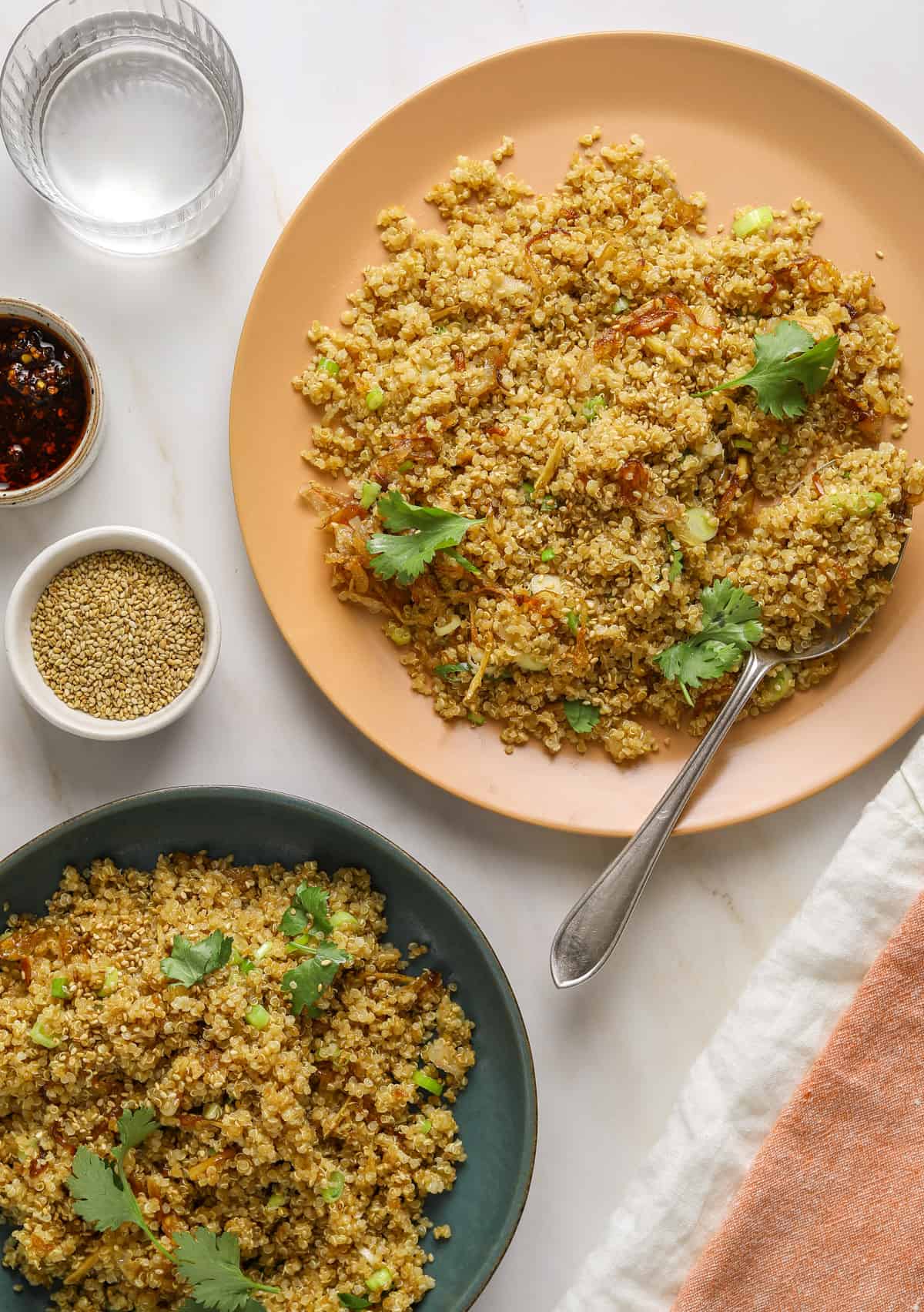 a pink plate filled with quinoa fried rice with a dish of chili oil and a bowl of sesame seeds