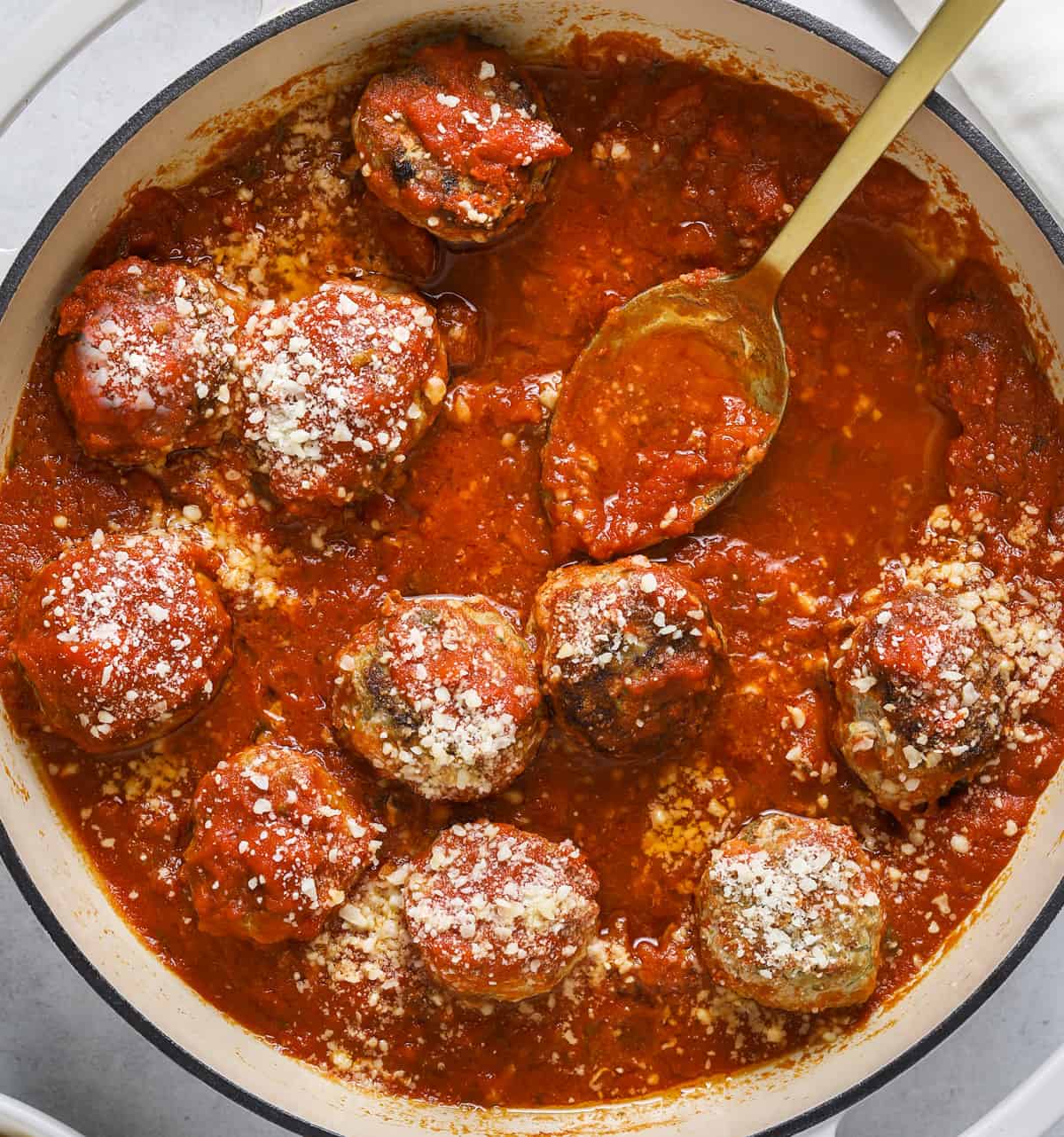 A close up image of turkey meatballs in a pot with red tomato sauce and grated parmesan cheese and a gold spoon