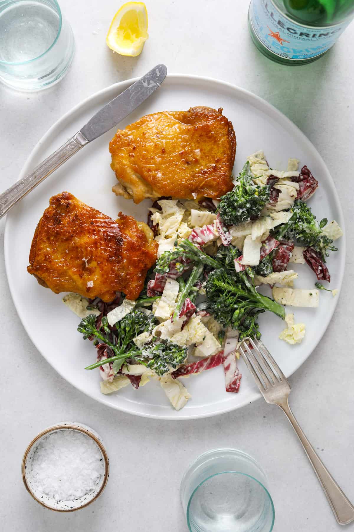 a white plate filled with two golden brown chicken thighs and a broccoli, radicchio slaw.