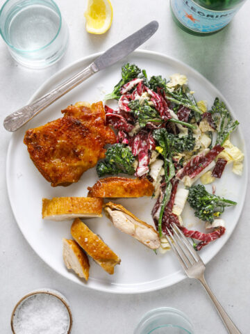 a round white plate with crispy chicken thighs and broccolini slaw. a bottle of San Pellegrino. two light blue glasses of water on the side.