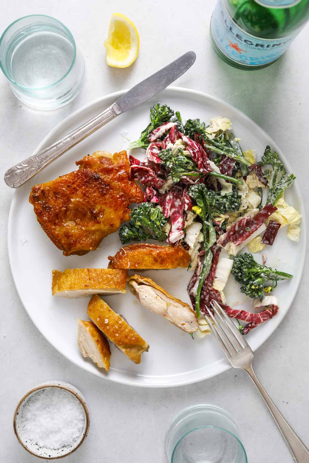 a round white plate with crispy chicken thighs and broccolini slaw. a bottle of San Pellegrino. two light blue glasses of water on the side.