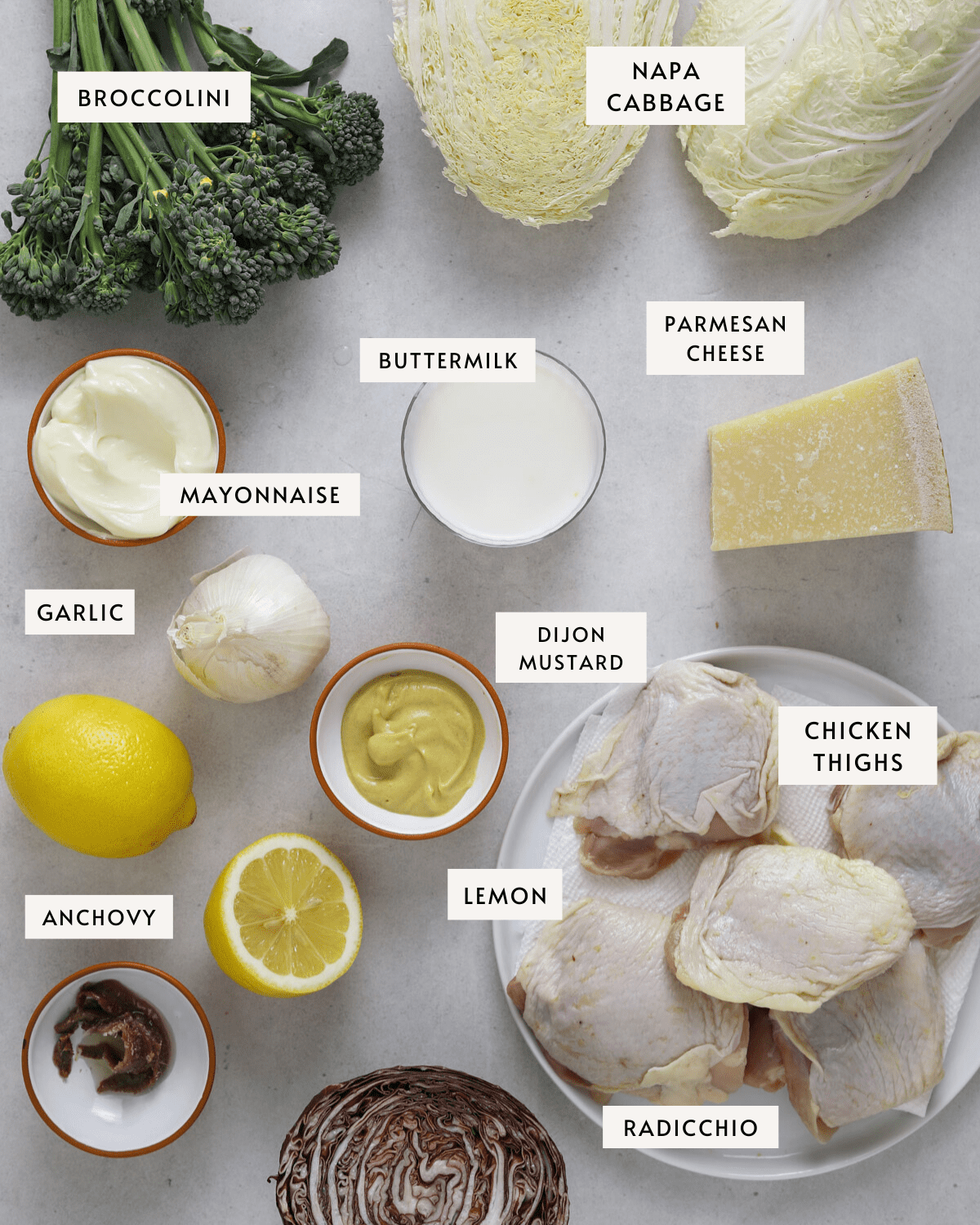 a bunch of broccolini, two halves of a head of napa cabbage, a bowl of dijon mustard, two lemons, chicken thighs of a white plate, radicchio, a bowl of buttermilk, a wedge of parmesan cheese.