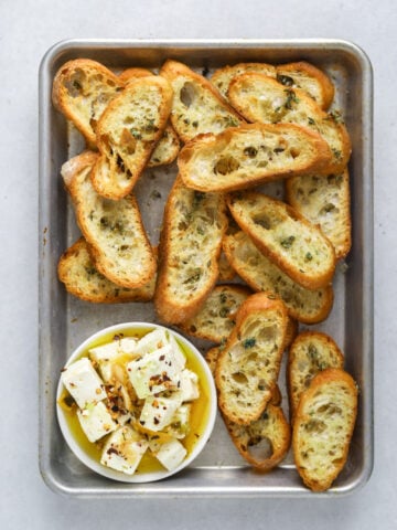a baking tray with toasted crostini and a small dish of marinated feta cheese