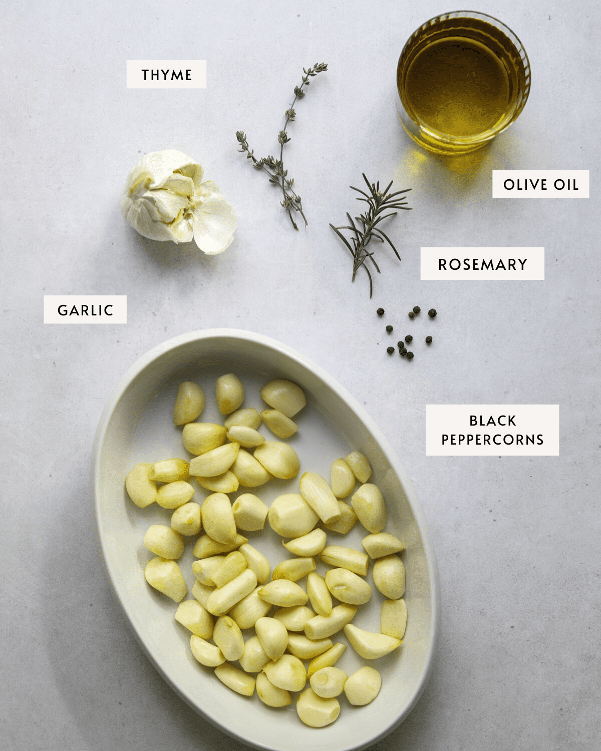 a white oval baking dish filled with raw garlic cloves, a whole head of garlic, black peppercorns, a glass dish of olive oil, sprigs of fresh rosemary and thyme