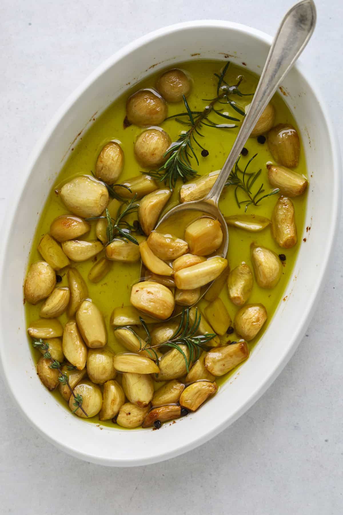 a white oval baking dish filled with roasted garlic cloves, olive oil and rosemary