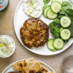 a white plate with a turkey and zucchini burger, spinach, sliced cucumber and a dollop of tzatziki