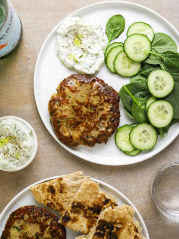 a white plate with a turkey and zucchini burger, spinach, sliced cucumber and a dollop of tzatziki
