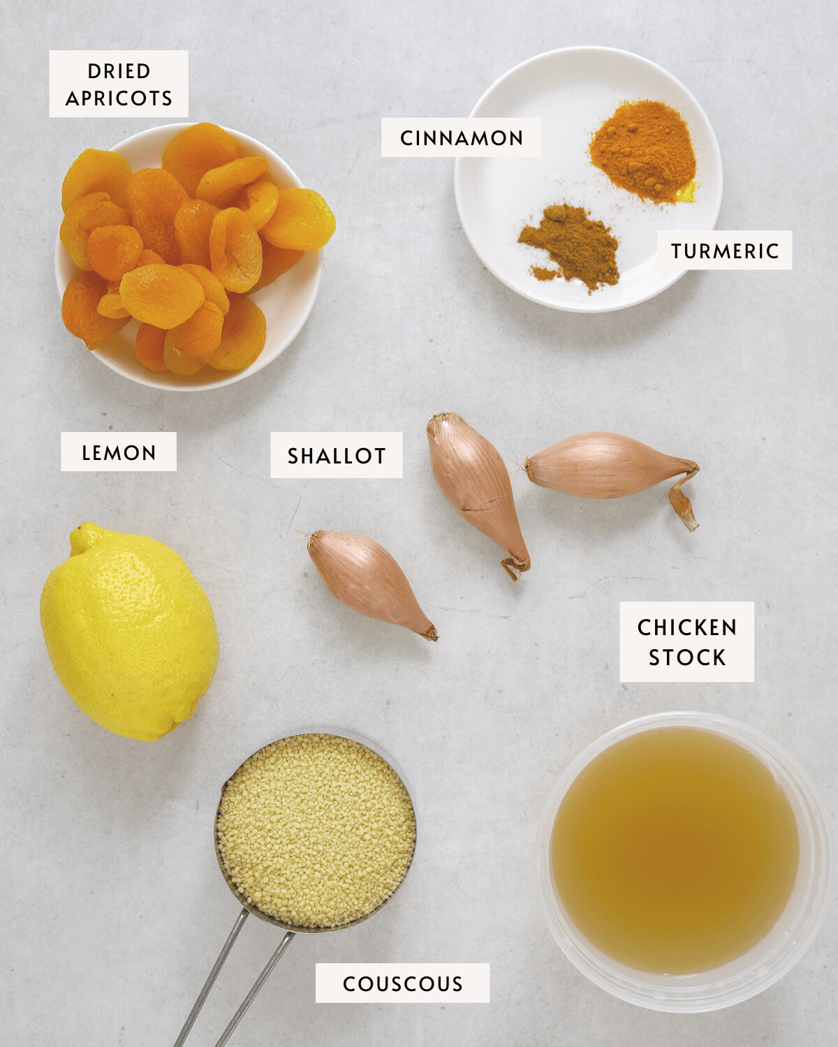 a small white bowl of dried apricots, a white bowl of cinnamon and turmeric, a container of chicken stock, three shallots, a lemon and a measuring cup of couscous