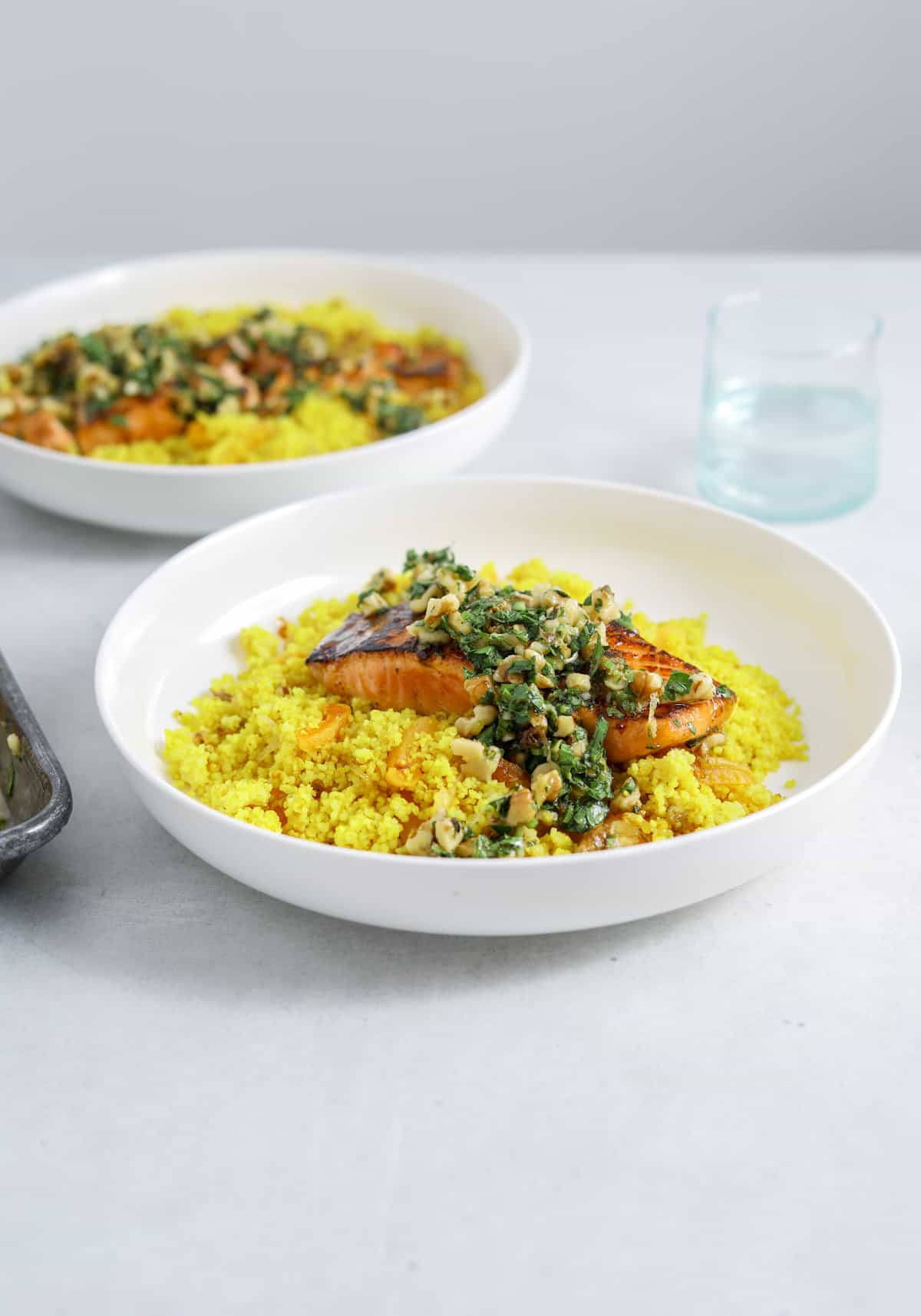 A bowl of yellow couscous in a bowl topped with a filet of salmon and chopped herb sauce on a blue table with a second bowl of salmon in the background