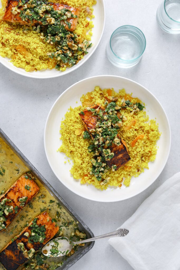 two white bowls of couscous and salmon with a baking tray of salmon filets off to the side