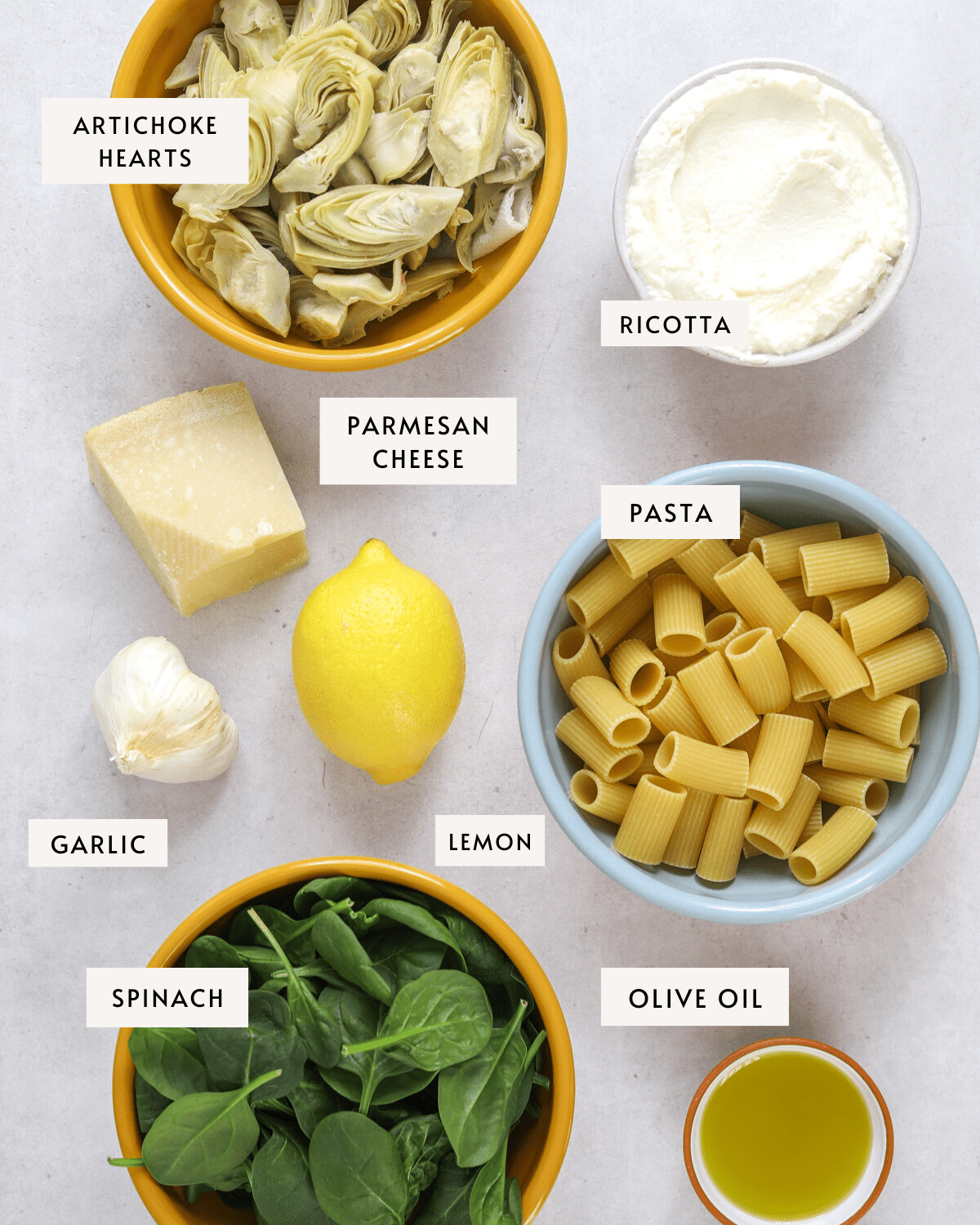 Recipe ingredients portioned in bowls: frozen artichoke hearts, ricotta cheese, rigatoni pasta, baby spinach, olive oil, a lemon, a head of garlic and a hunk of parmesan cheese.