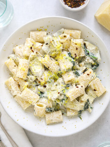 a white speckled ceramic bowl filled with creamy ricotta pasta, lemon zest and wilted spinach.