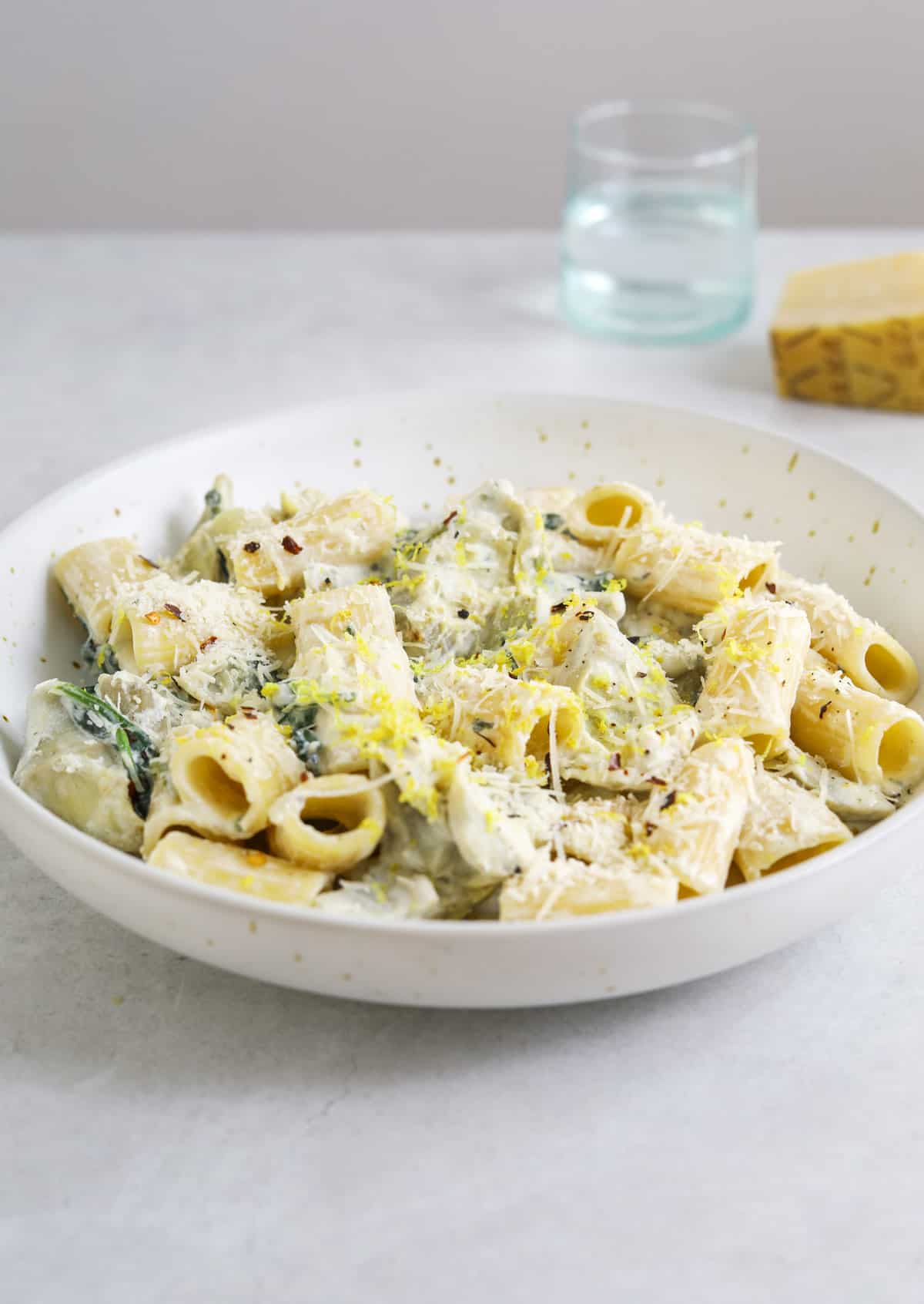 a white bowl filled with creamy ricotta pasta on a blue background with a light blue glass and a hunk of parmesan cheese in the background.