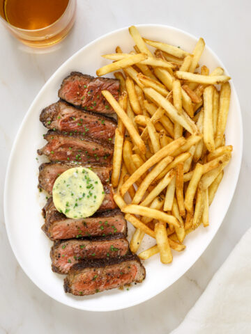 a white oval platter filled with sliced steak, a disk of blue cheese compound butter, french fries and a cocktail on the side