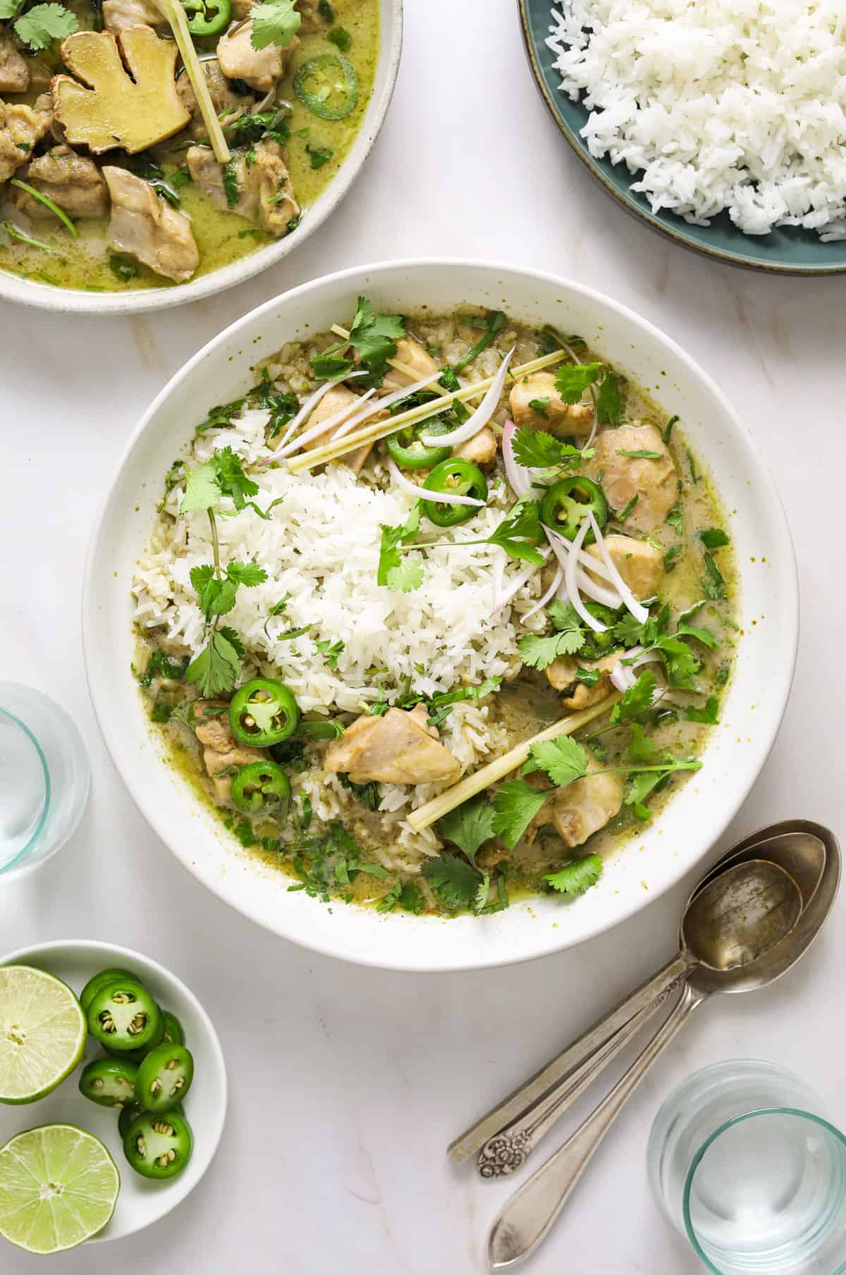 a white bowl filled with rice, green coconut curry sauce, chicken, cilantro, jalapeño slices and lemongrass.