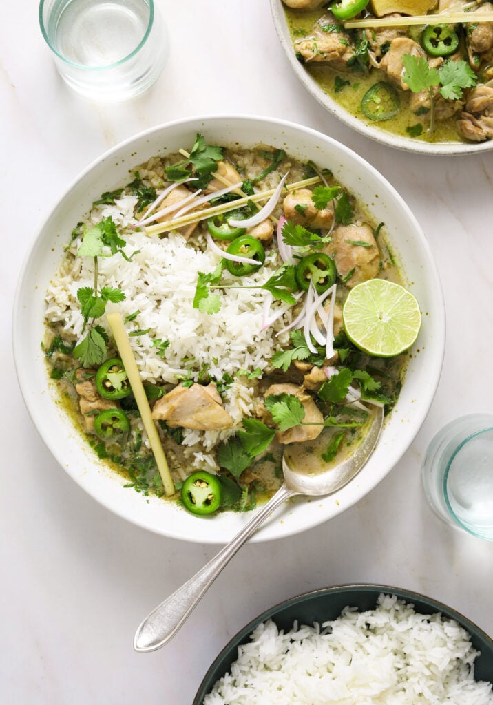 a white ceramic bowl filled with green chicken curry. two glasses of water and a bowl of rice.