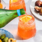 a bottle of sparkling water being poured into a clear glass filled with an Aperol spritz