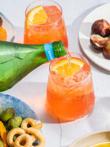 a bottle of sparkling water being poured into a clear glass filled with an Aperol spritz