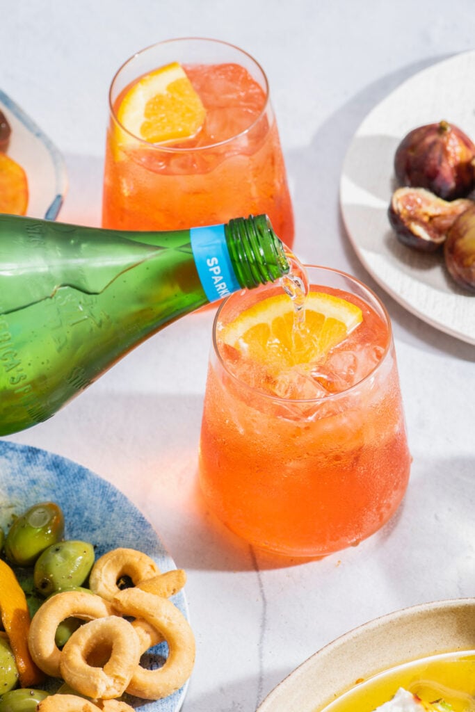 a bottle of sparkling water being poured into a clear glass filled with an Aperol spritz