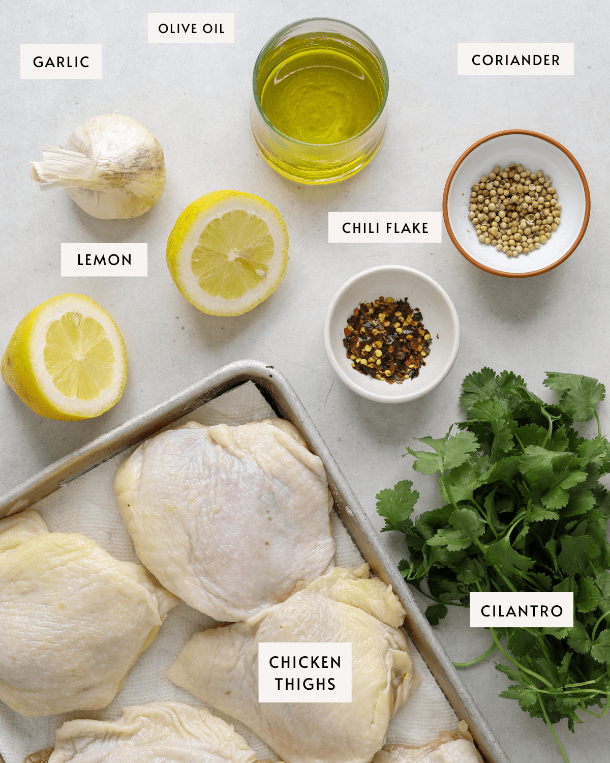 a baking tray with raw chicken thighs, tiny bowls of coriander and chili flakes, a lemon sliced in half, a bulb of garlic and a bunch of cilantro.