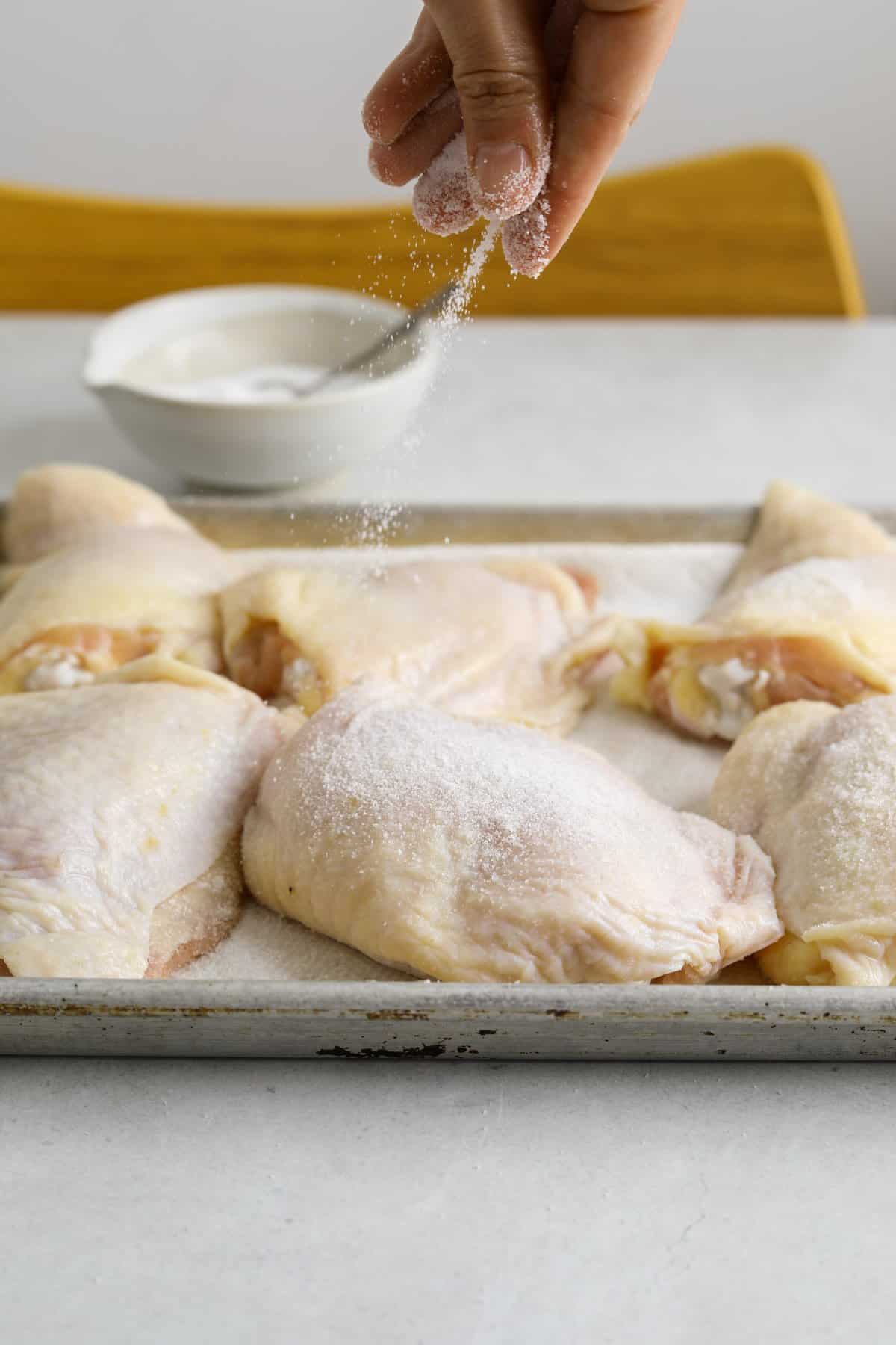a baking tray with chicken thighs being seasoned with salt on a blue table top.