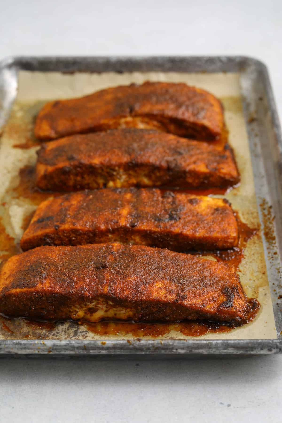 Four blackened oven-roasted bbq salmon filets on a parchment lined baking tray.
