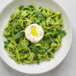 a white speckled ceramic bowl filled with bright green pea pesto pasta and a dollop of creme fraiche drizzled with olive oil.