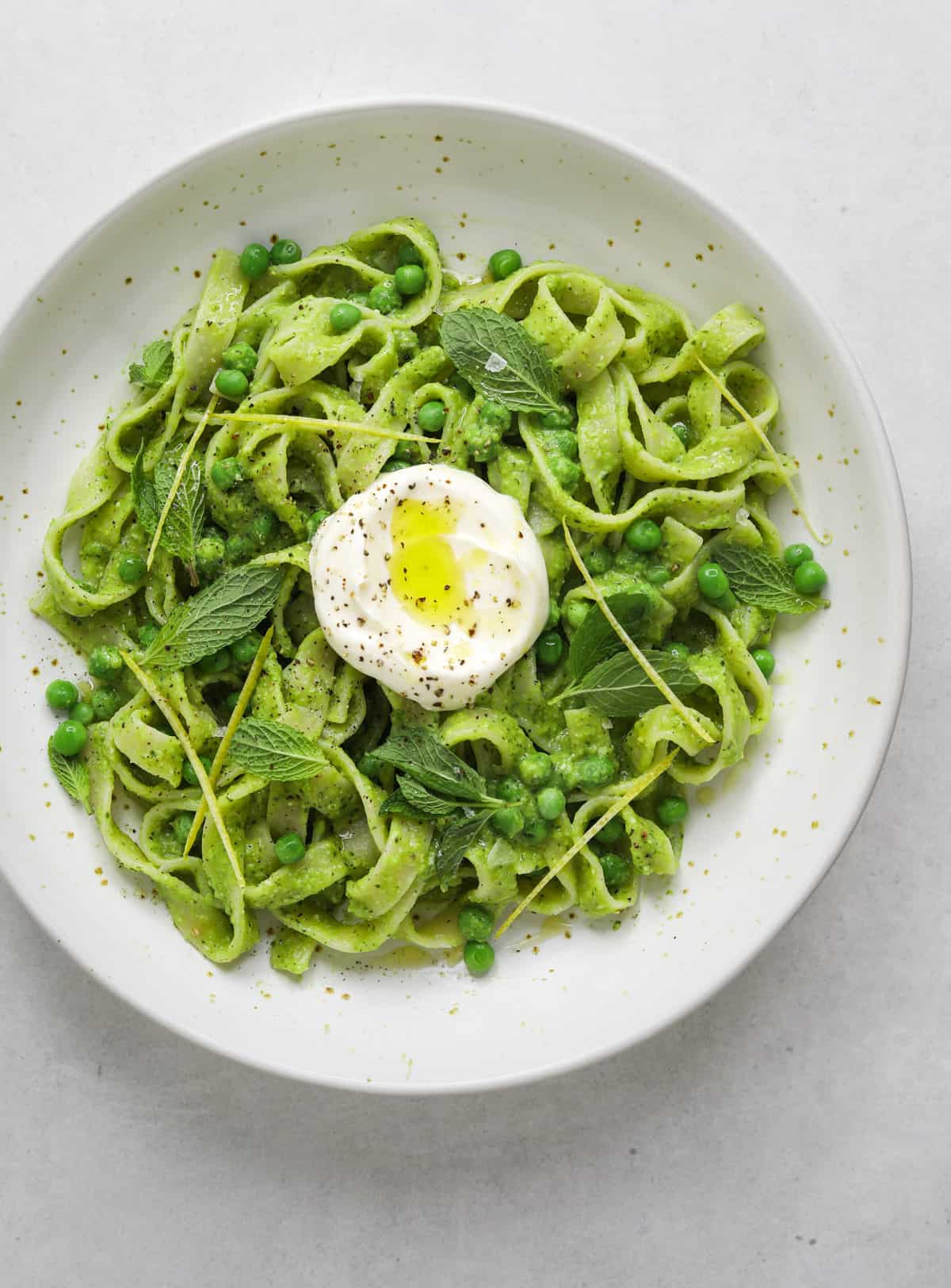 a white speckled ceramic bowl filled with bright green pea pesto pasta and a dollop of creme fraiche drizzled with olive oil.