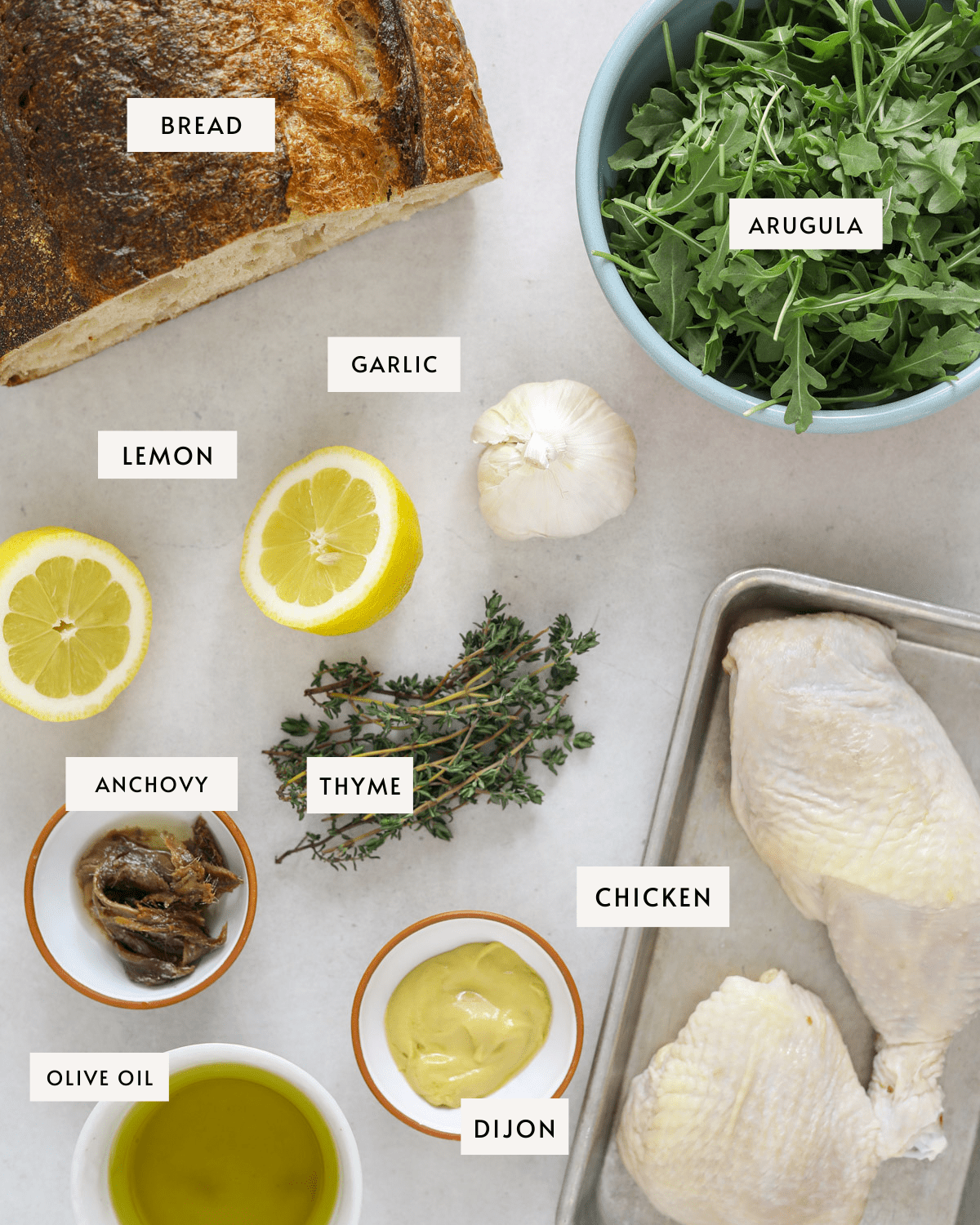 recipe ingredients portioned on a light grey backdrop: a loaf of bread, a bowl of arugula, raw chicken, a bunch of fresh thyme, a head of garlic, lemon, and olive oil.