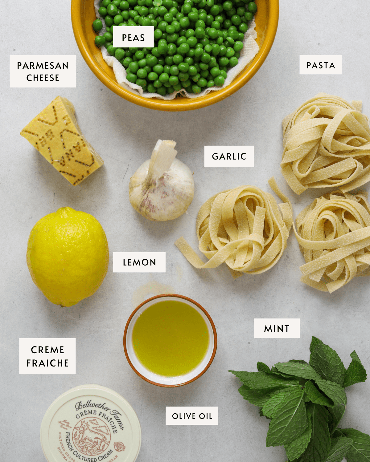 recipe ingredients individually portioned: frozen peas in a yellow bowl, a lemon, a head of garlic, tagliatelle pasta, fresh mint, creme fraiche and olive oil.
