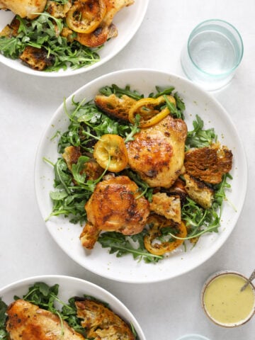 A white bowl filled with arugula salad, roast chicken, croutons and roasted lemon rounds. A small bowl of vinaigrette on the side as well as a small blue glass of water.