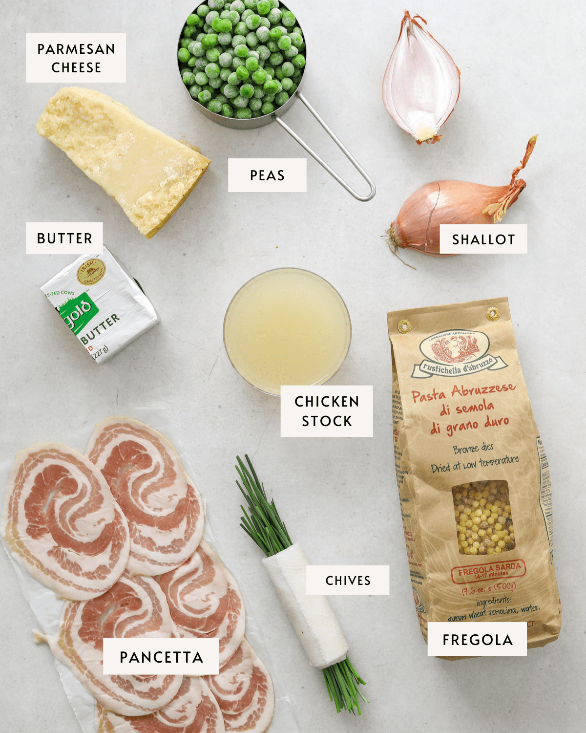 a hunk of parmesan cheese, raw sliced pancetta, a bundle of chives, a bag of Fregola, a shallot sliced in half, a small glass bowl of chicken stock