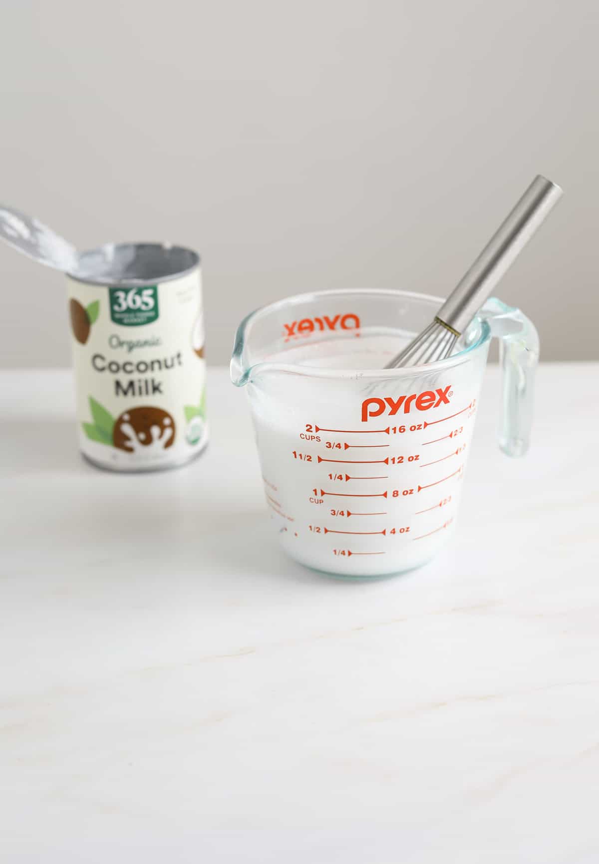 A glass Pyrex measuring cup with a whisk and a can of coconut milk in the background.