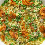 a white bowl filled with fregola, chicken broth, chives, grated parmesan cheese and crispy bits of pancetta