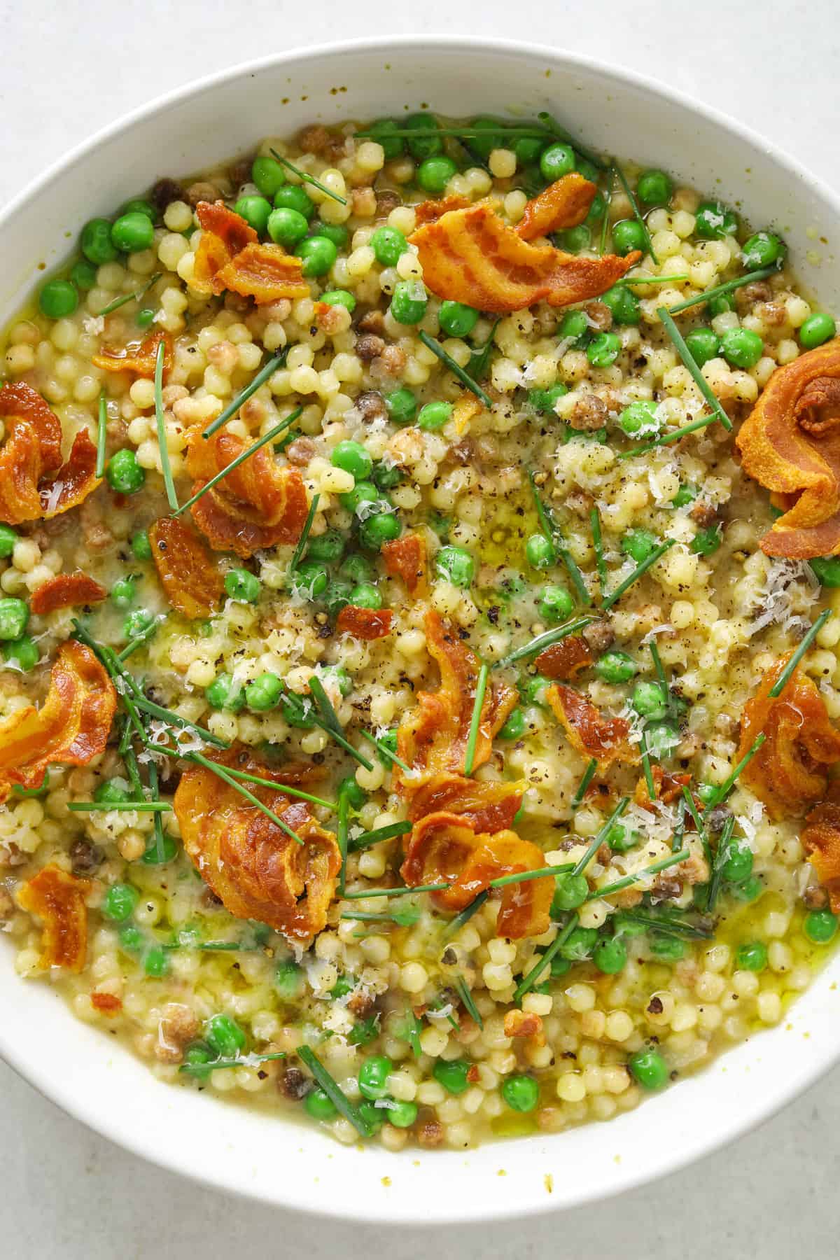 a white bowl filled with fregola, chicken broth, chives, grated parmesan cheese and crispy bits of pancetta