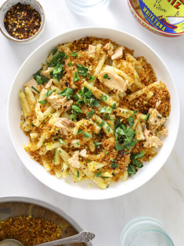 a white bowl with pasta surrounded by a glass of water, a bowl of breadcrumbs, a can of tuna and a small dish of chili flakes