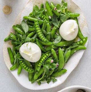 a white and beige platter filled with snap pea salad, pea tendrils and two balls of fresh burrata cheese.