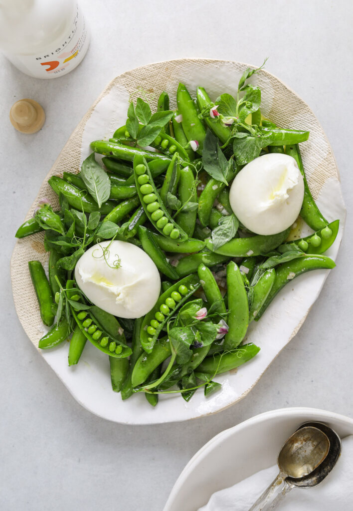 a white and beige platter filled with snap pea salad, pea tendrils and two balls of fresh burrata cheese.