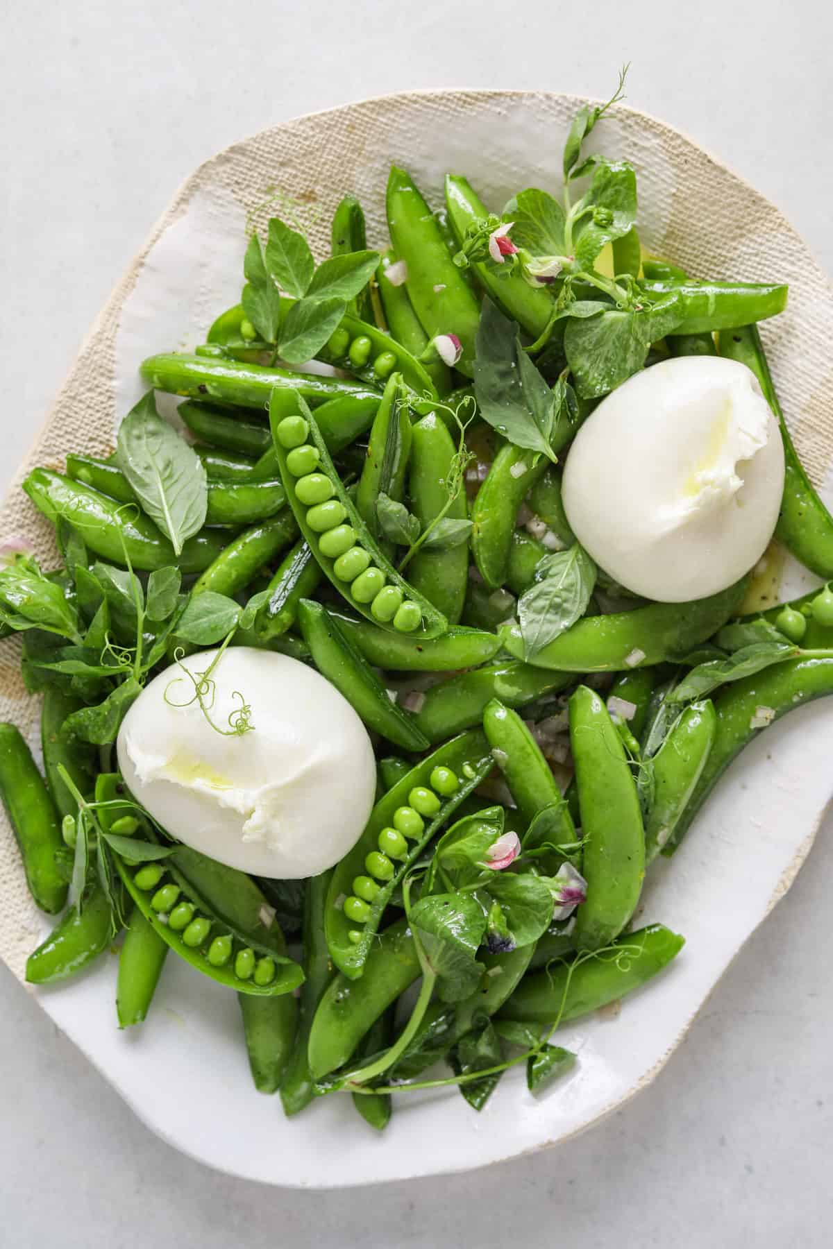 a close up photo of a snap pea and burrata salad on a light blue/grey background.