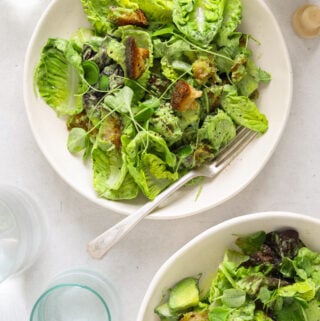 two white bowls full of lettuce, croutons, herbs and avocado. Three water glasses and a bottle of olive oil.