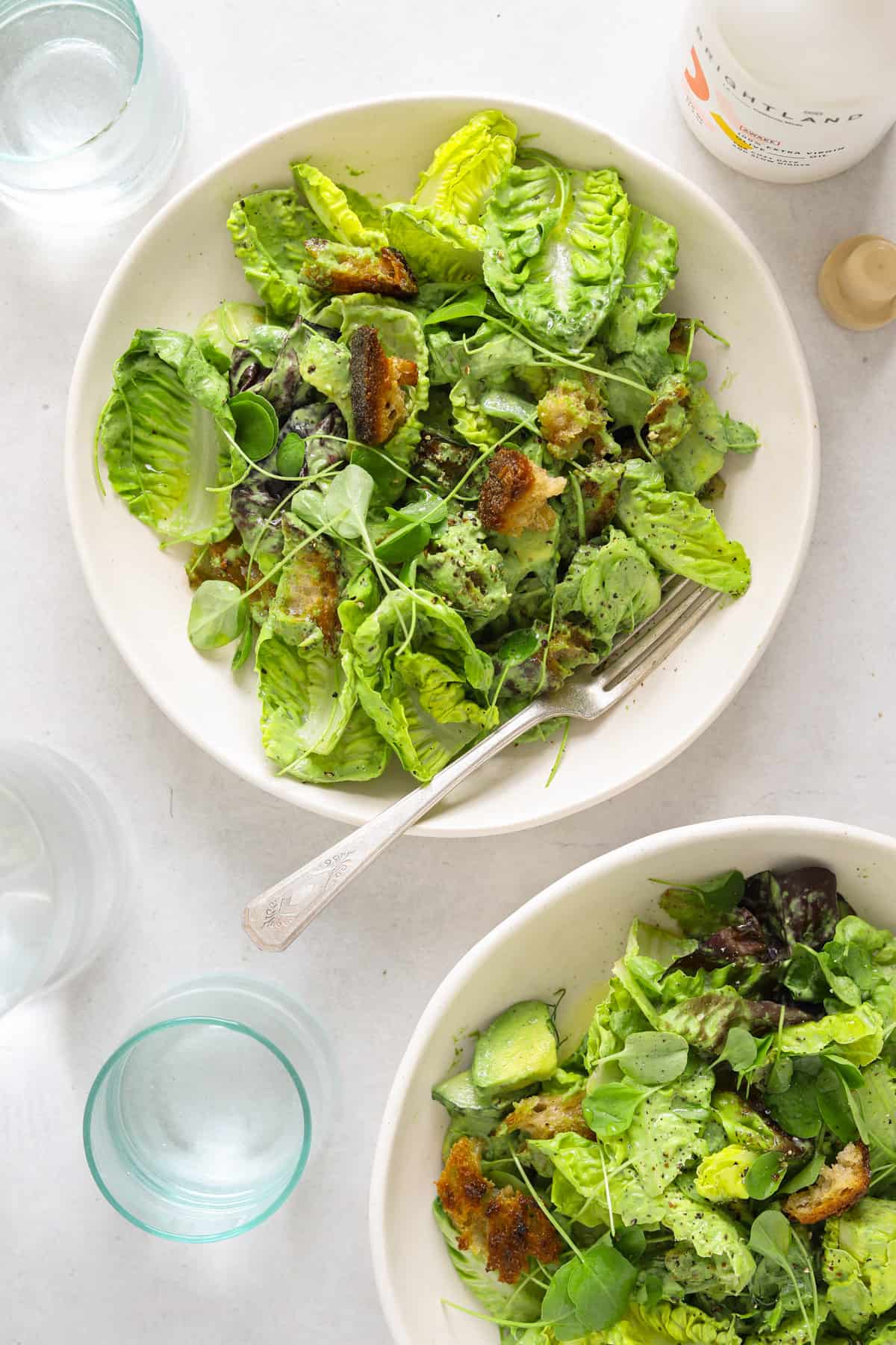 two white bowls full of lettuce, croutons, herbs and avocado. Three water glasses and a bottle of olive oil.