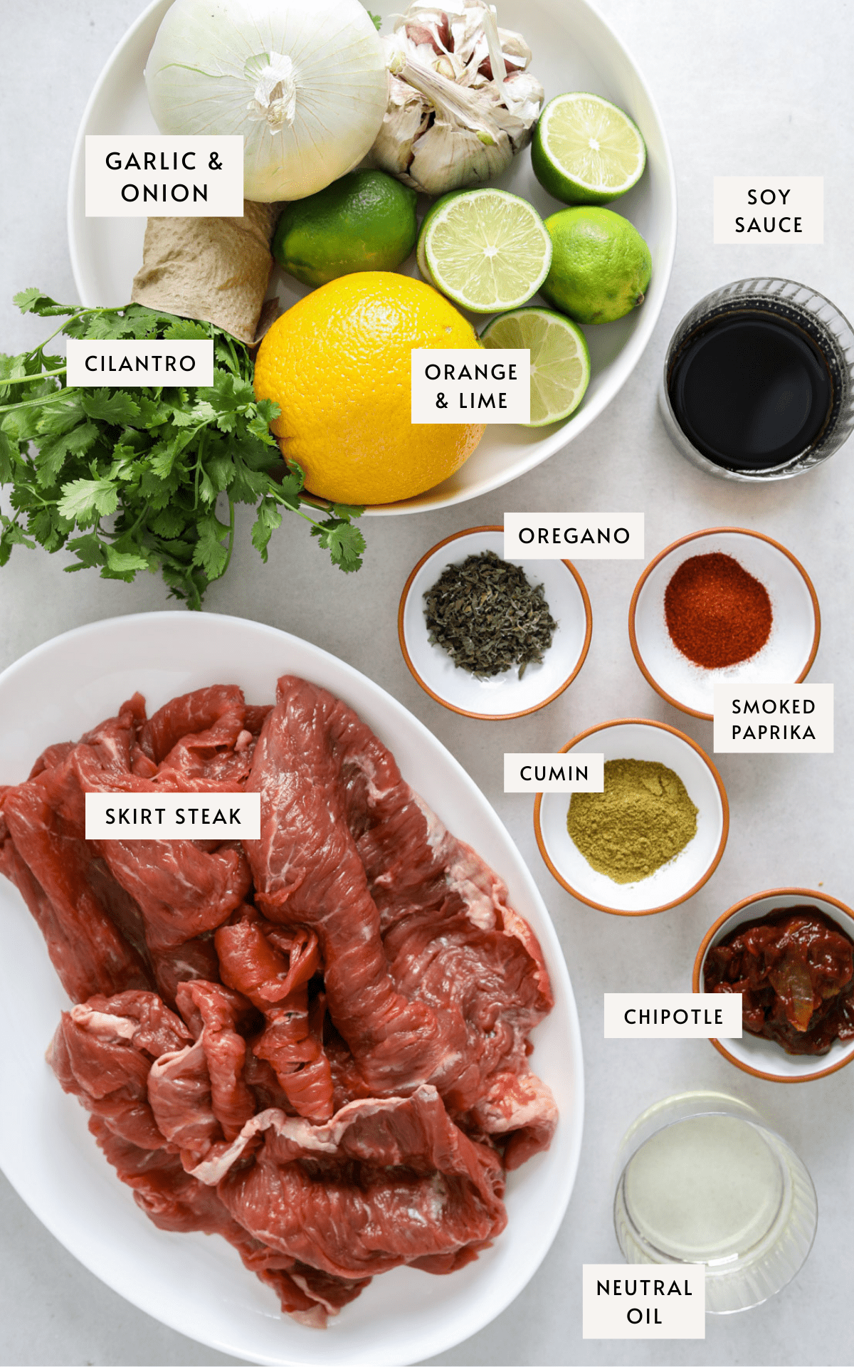 a white oval platter with raw skirt steak, small bowls filled with spices, a large bowl with oranges, limes, garlic bulbs, onion and cilantro, chipotle peppers in adobo sauce and a small glass bowl of neutral oil.