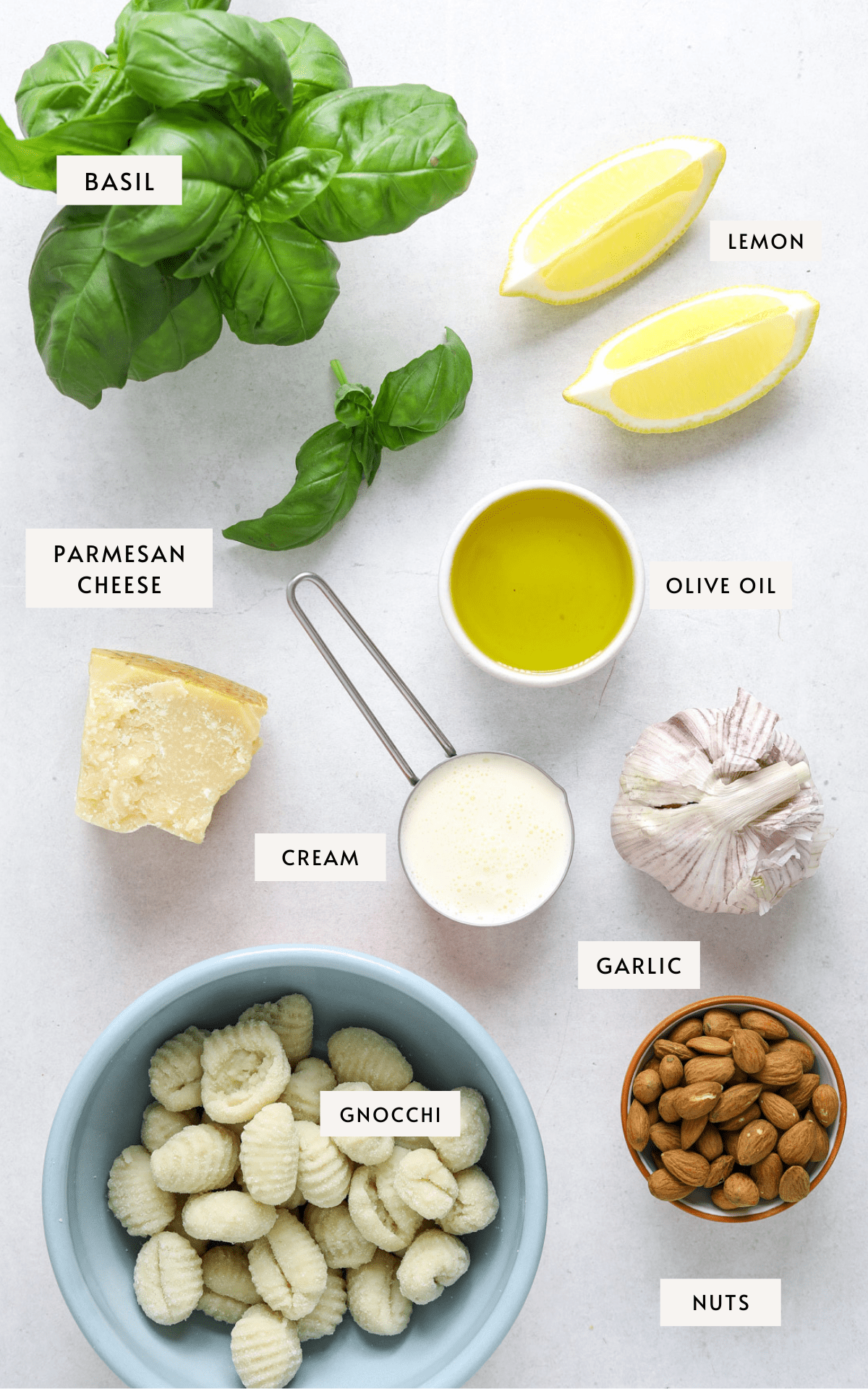 Fresh basil leaves, two lemon wedges, parmesan cheese, cream in a measuring cup, garlic, olive oil, a small bowl of almonds, gnocchi in a blue bowl.