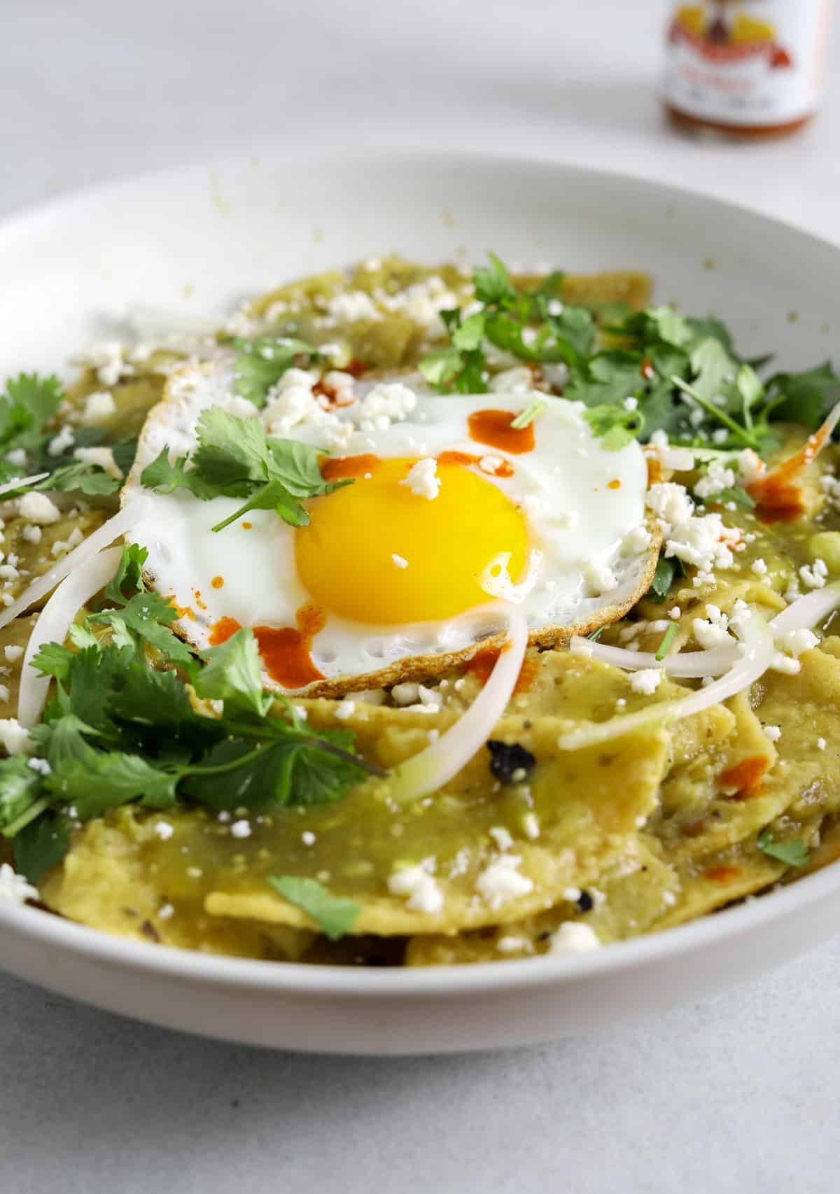 A white ceramic bowl filled with chilaquiles, salsa verde, cilantro, crumbled cheese and a sunny side up egg.
