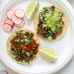 Two tacos, two lime wedges and sliced radishes on a white paper plate surrounded by Mexican sodas and cups of salsa.