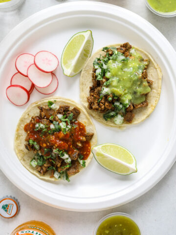 Two tacos, two lime wedges and sliced radishes on a white paper plate surrounded by Mexican sodas and cups of salsa.