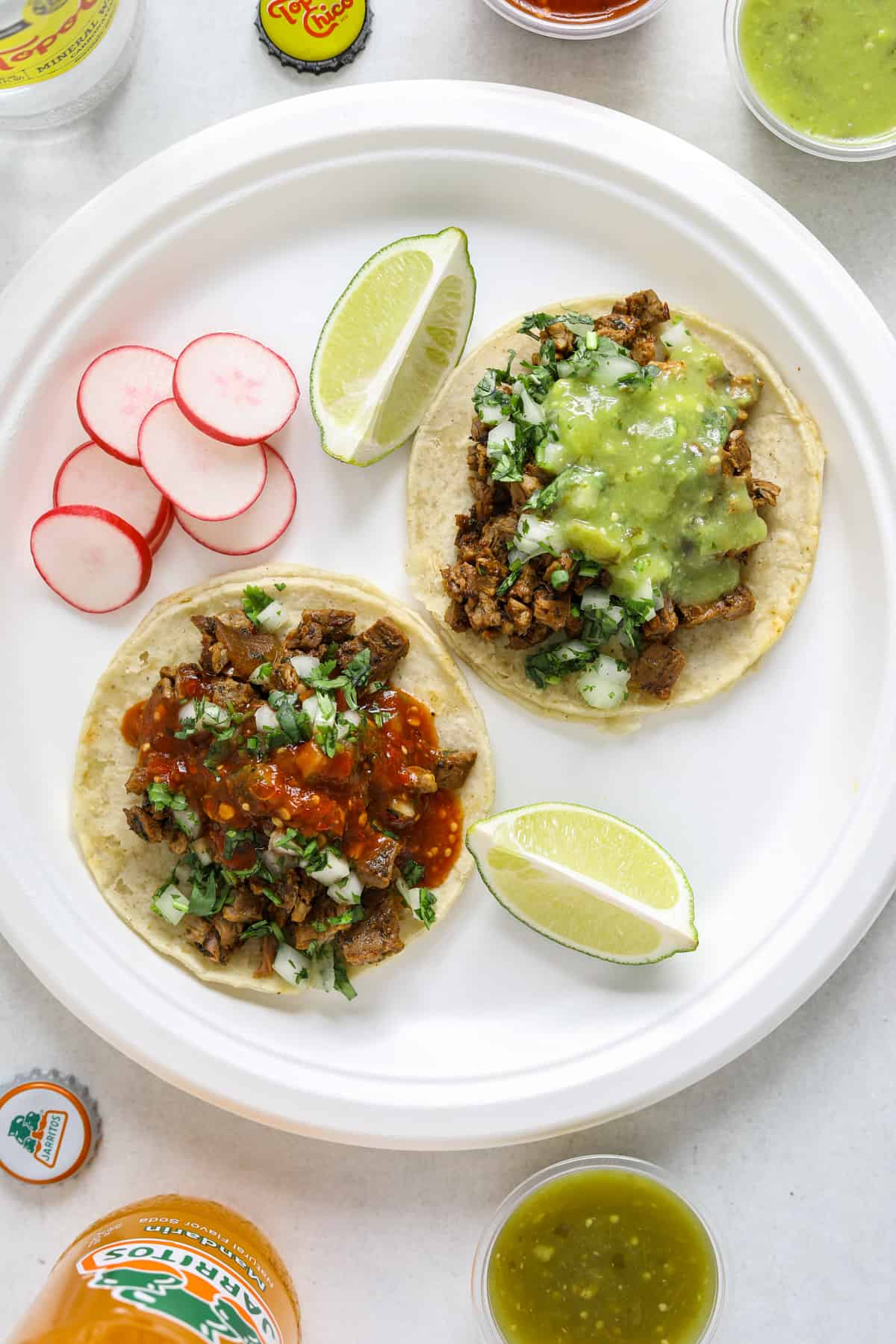 Two tacos, two lime wedges and sliced radishes on a white paper plate surrounded by Mexican sodas and cups of salsa.