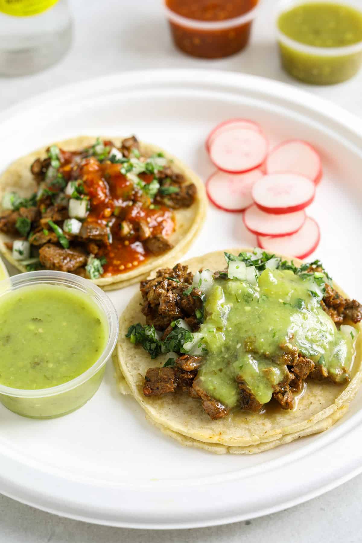 A paper plate filled with two tacos, sliced radish and a small cup of salsa verde.