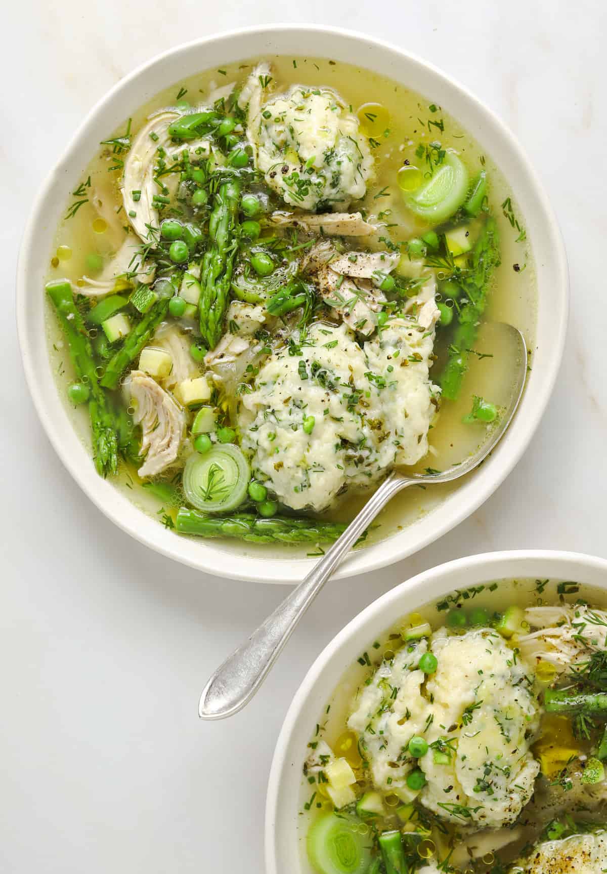 a white bowl filled with chicken soup and herb dumplings, asparagus, peas and leeks.